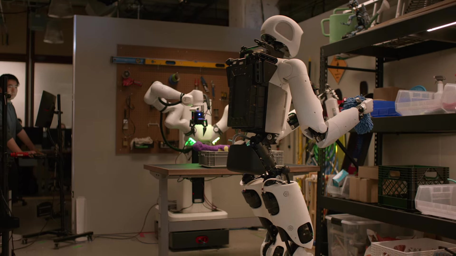 An Apollo humanoid carries a black crate while a stationary robot arm works at a nearby table