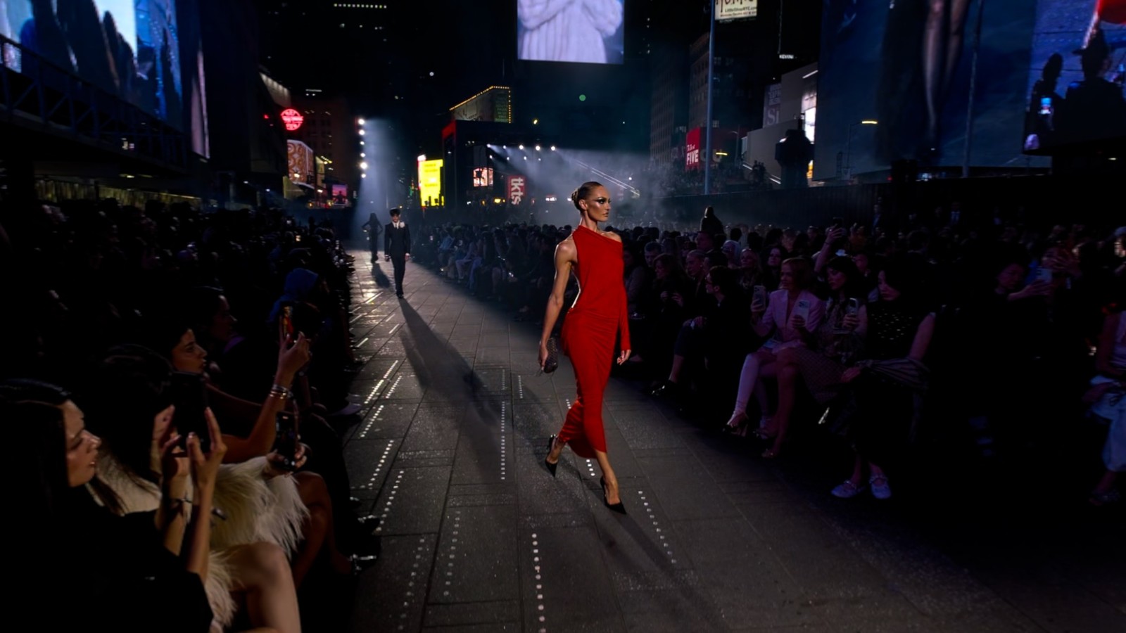 A model in a red dress walks between guests at Gucci’s GucciCore show in Times Square, shown in an official Gucci app screenshot