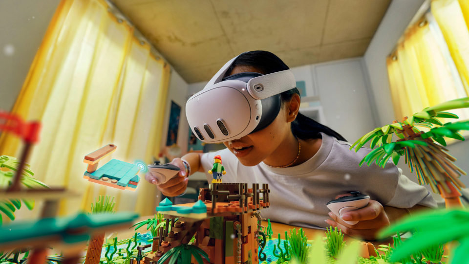 A person wearing Meta Quest 3 and holding controllers as mixed reality game pieces fill the room