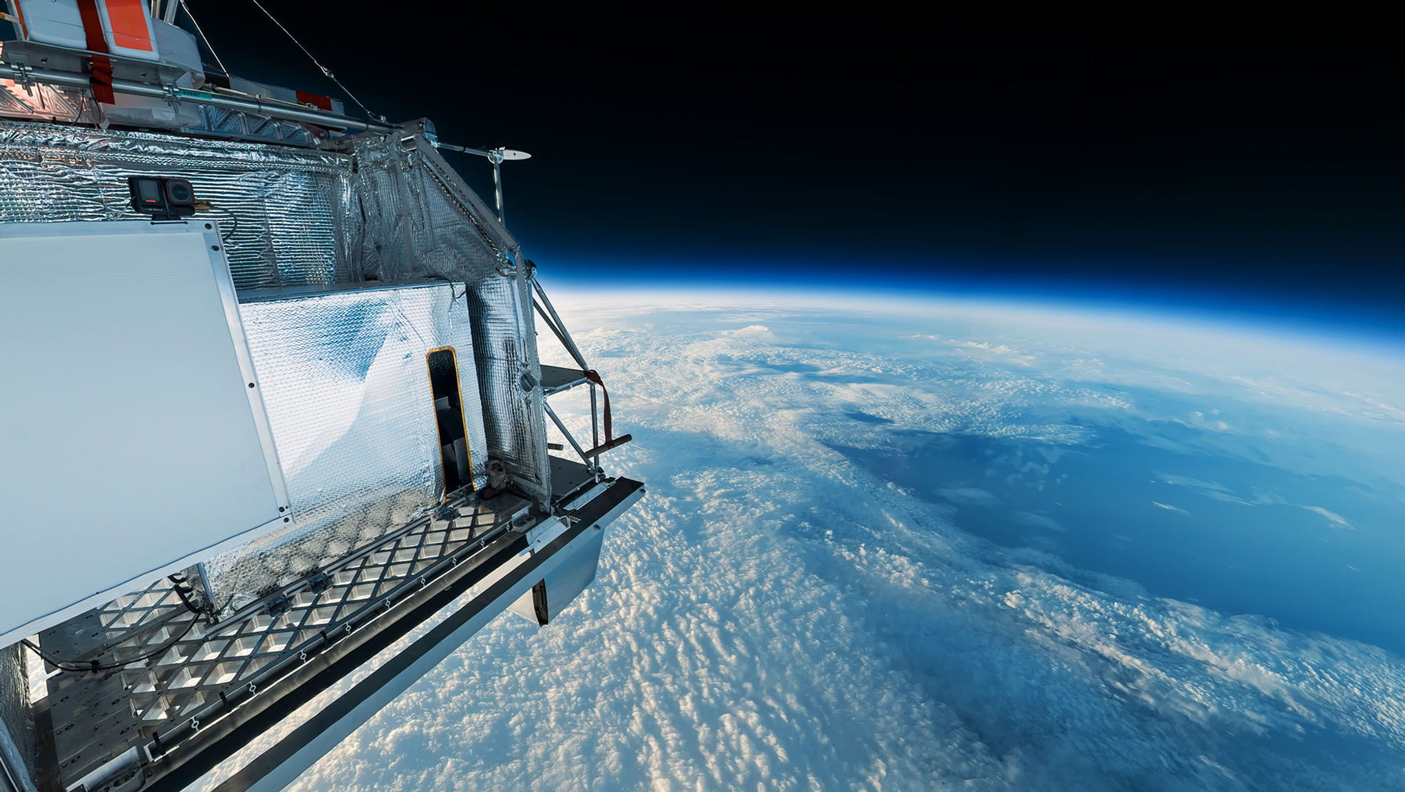 The STRATOS balloon gondola above clouds with Earth’s curved horizon and a thin blue atmosphere against black sky