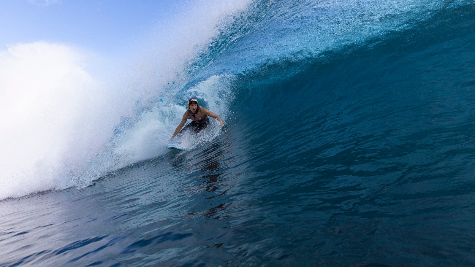 A surfer rides beneath a large curling blue wave at Teahupoʻo, Tahiti.