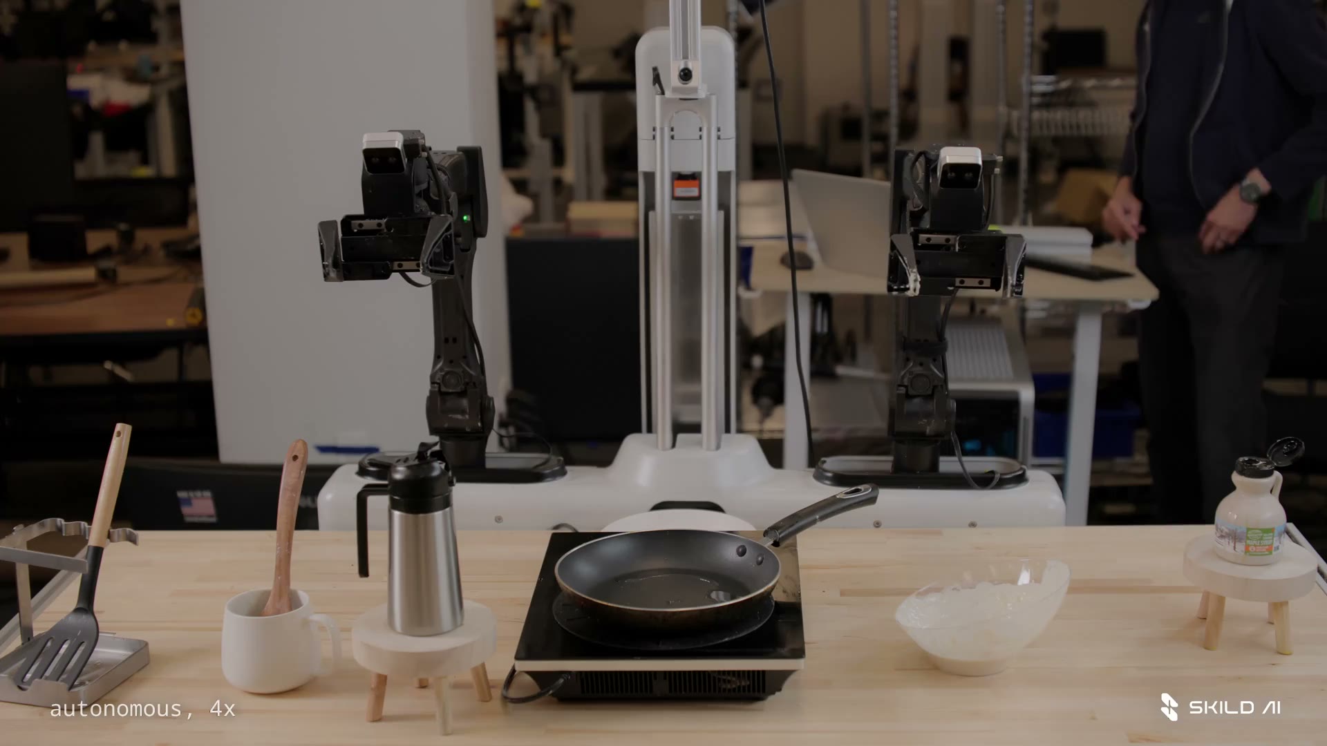 A two-armed robot stands behind a pan and pancake ingredients